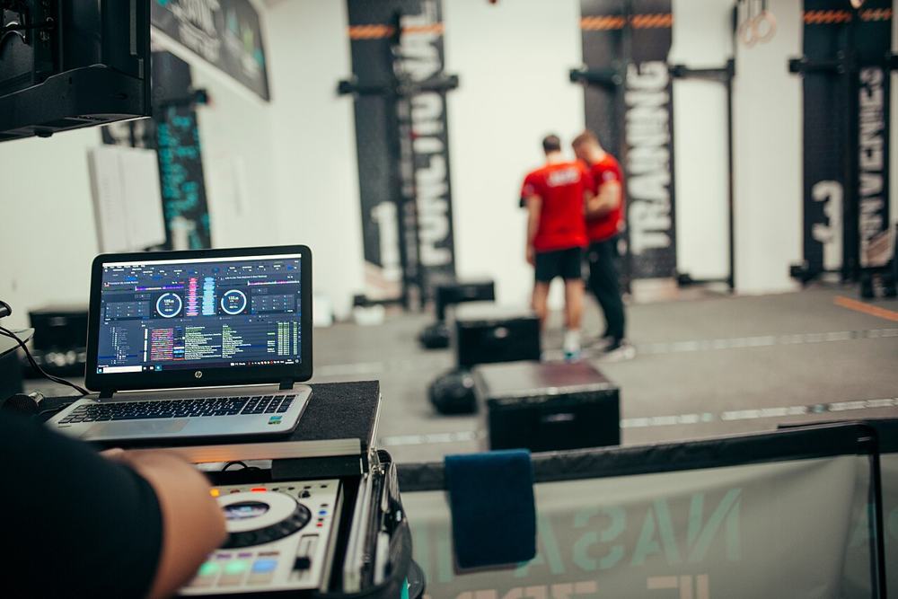 A DJ operates a laptop with music software while two fitness instructors discuss their training program in a well-equipped gym.jpg